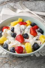Delicious cheesecake fruit and berry salad with strawberries, blueberries, pineapple close-up in a bowl on a marble table. Vertical