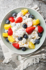 Delicious cheesecake fruit and berry salad with strawberries, blueberries, pineapple close-up in a bowl on a marble table. Vertical top view from above