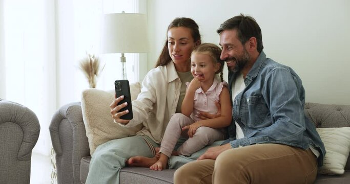 Loving parents with little 4s daughter using smart phone on weekend at home, sit on couch look at gadget screen, making videocall enjoy remote talk to family, having fun enjoy new mobile application