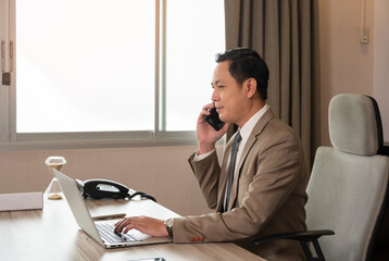 Busy Asian businessman or entrepreneur in brown suit talking on cellphone with client and working on laptop while sitting at his working place in personal office. - 777992639