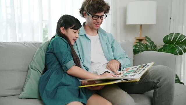 Caucasian father and cute daughter reading story book together while sitting at sofa. Happy dad telling story to cute american child to imagination development while having time together. Pedagogy.