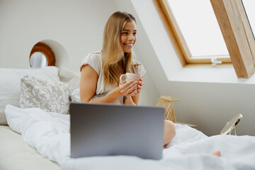 A woman is leisurely sitting on a wooden bed, with a laptop on her arm and a cup of coffee in hand - obrazy, fototapety, plakaty