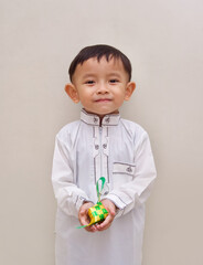  A cute little boy from Asia wearing a white Islamic long shirt with a simple design, holding a small green and yellow colored Ketupat in his hand, with a smiling facial expression