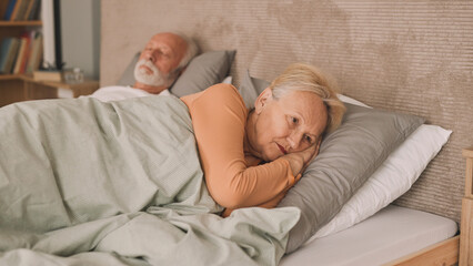 Sleepless elderly lady lying in bed next to her husband