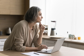 Thoughtful serious middle-aged woman work at home with laptop, look away, touch chin in deep...