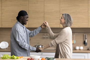 Happy middle-aged multiethnic affectionate couple enjoy romantic date, distracted from dish preparation, holding hands, moving to favourite music, dancing in modern kitchen. Relations, leisure, love