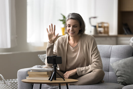 Smiling attractive mature woman sit on sofa record videoblog using smart phone camera, greeting subscribers, start live on-line stream event. Webinar, online counselling, vlogging, social media icon