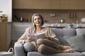 Middle-aged woman rest at home with favorite book, sit on sofa, enjoy weekend alone in living room,...
