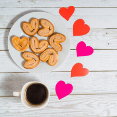 A cup of coffee, a plate with heart-shaped cookies on a wooden white table. Background with...