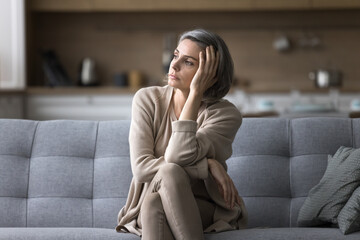 Attractive silent middle-aged woman relaxing seated on couch in living room, staring out window, ponders, missing, deep in thoughts, looks pensive and thoughtful, spend boring weekend alone at home - obrazy, fototapety, plakaty