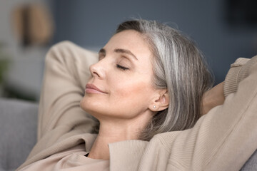 Close up peaceful mature female relaxing with eyes closed in living room sitting on sofa, breath...