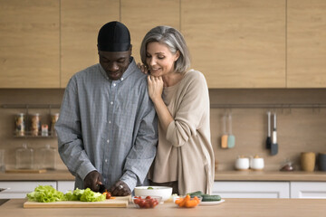 Positive couple in love cooking together healthy dish, cutting tomatoes, lettuce for vegetarian salad on weekend at home. Multiethnic spouses enjoy cookery process and communication in modern kitchen