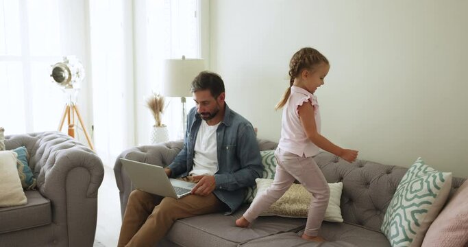 Focused middle-aged man sitting on couch working on wireless computer, do business task, engrossed on remote job, his little 4s daughter disturbing daddy, jumping nearby. Telework, freelance from home