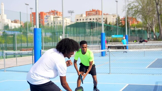 Side view full length video of two african american young male friends having fun during pickleball match outdoors