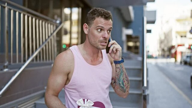 Handsome man with tattoos holds gift, talks on phone in urban setting.