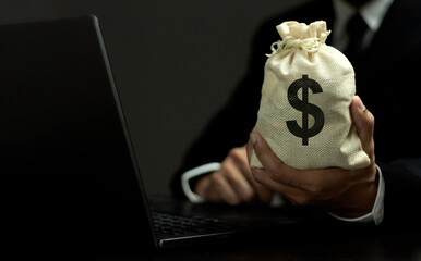 A person is holding a bag of money in front of a laptop