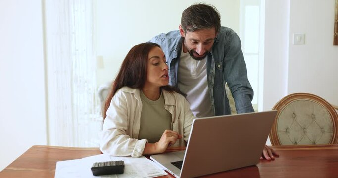 Pretty Portuguese spouses review bank statements displayed on laptop screen, use e-bank system to track expenses, perform incomes calculations. Savings, family budget, finances planning and management