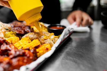 A close-up view of a tray of food with mustard being drizzled on grilled sausages and corn, showcasing a delicious and appetizing meal