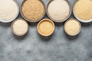 Border of different varieties of dry rice in bowls on a gray grunge background. Top view, copy space.