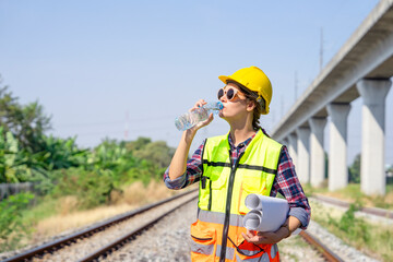 railway engineer or construction worker in hardhat and safety vest,holding paperwork is thirsty in the harsh sunlight,attractive caucasian female wears sunglasses drinking water while working outside - obrazy, fototapety, plakaty