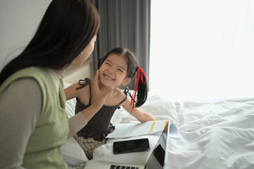 Mother using laptop working distantly from home and playing with little daughter in cozy bedroom