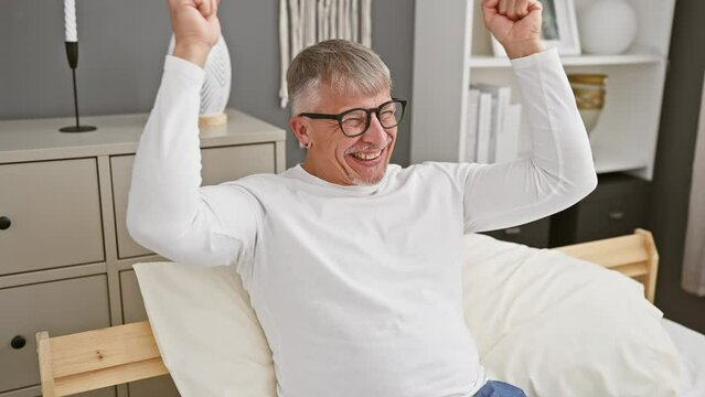 Joyful middle age, grey-haired man, celebrating a triumphant win with arms raised in victorious excitement, jubilantly shouting 'yes' from his homely bedroom, wearing his winning pyjamas.