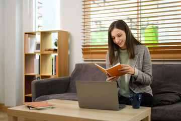 Smiling caucasian woman reading notes and working with laptop in living room