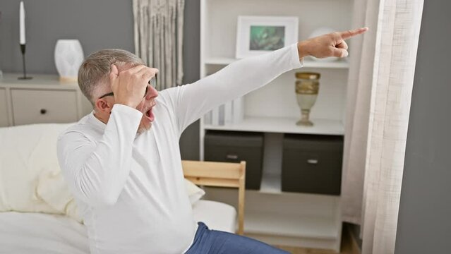 Amazed middle age grey-haired man in pyjamas, open mouthed surprise, pointing ahead at something astounding in his bedroom.