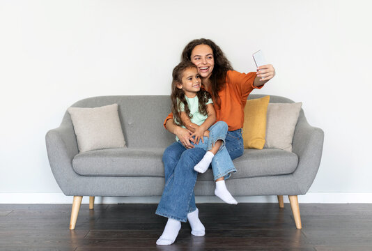 Cheerful mother and little girl taking selfie sitting on couch, family spending time together, happy mom and daughter making video call