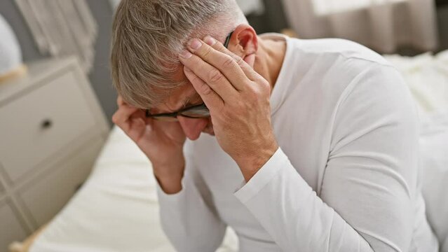 Stressed middle age grey-haired man suffering from a head-splitting migraine in bedroom, hand wringing in pain, the ache showing on his face, wearing pyjamas.