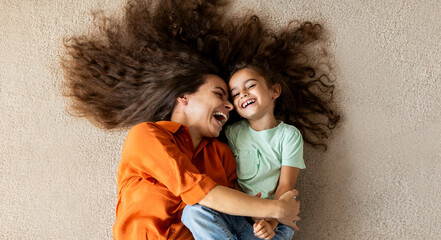 Top view of happy mother and her little daughter lying on soft carpet, laughing, having fun at...