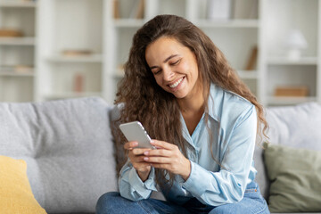 Cheerful young european lady typing on smartphone, resting in living room interior, using social media chat, app, reading good news