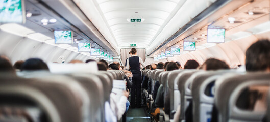 Interior of airplane with passengers on seats and stewardess in uniform walking the aisle, serving people. Commercial economy flight service concept - obrazy, fototapety, plakaty