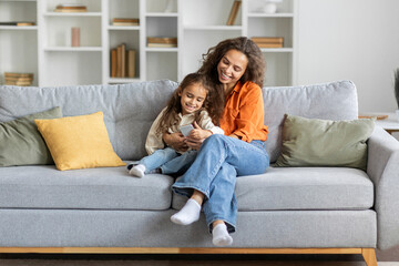 Smiling woman and little girl using cellphone, sitting on couch in living room, browsing internet,...
