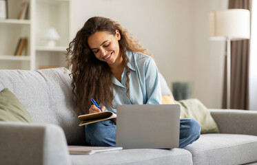 Happy female student writing notes while watching online class on laptop computer, sitting on sofa...