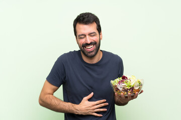 Young handsome man with salad over isolated green wall smiling a lot