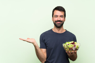 Young handsome man with salad over isolated green wall holding copyspace imaginary on the palm to...