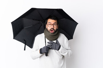 Caucasian handsome man with beard holding an umbrella over isolated white wall surprised and shocked while looking right