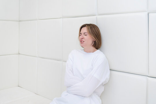 Young frustrated girl in a straitjacket is in an isolated white room in a mental hospital	