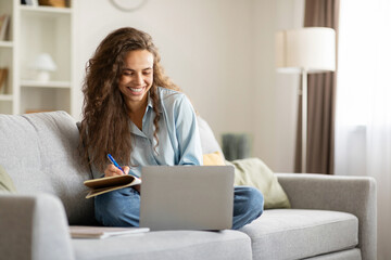 Smiling female student watching online class on laptop computer and writing notes, sitting on sofa at home. Remote distant learning concept