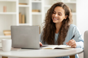 Positive cheerful young woman student using laptop and taking notes, attending webinar, have online...