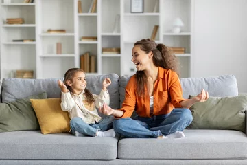 Poster Cute little girl with young mother sitting in lotus pose on couch at home, mum and daughter practicing yoga and laughing, having fun on weekend © Home-stock
