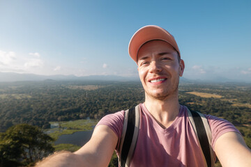 Happy man taking selfie photo from summer vacation day. Handsome tourist wearing cap and smiling at camera against tropical landscape.