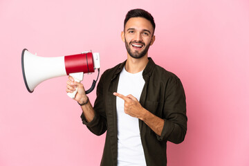 Young caucasian man isolated on pink background holding a megaphone and pointing side