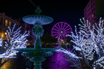 Grande roue illuminée en violet devant une fontaine la nuit à Noël