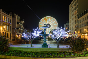 Grande roue illuminée en jaune devant une fontaine la nuit à Noël - obrazy, fototapety, plakaty