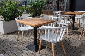 Outdoor cafe. Empty tables and chairs stand on cobblestone pavement next to green bushes in square concrete pots for street flowers