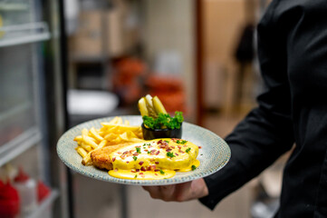 A waiter holds a plate of a delicious meal, featuring a creamy dish garnished with herbs and spices, accompanied by golden fries and fresh vegetables, showcasing culinary artistry