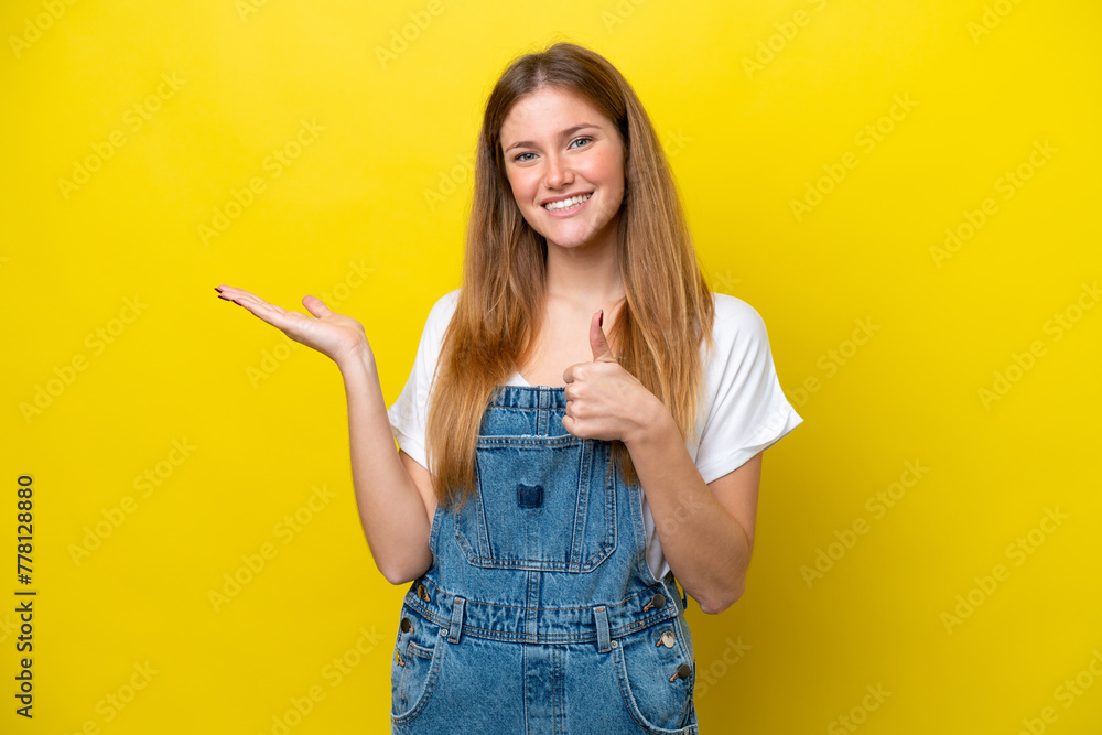 Wall mural Young caucasian woman isolated on yellow background holding copyspace imaginary on the palm to insert an ad and with thumbs up - Wall murals