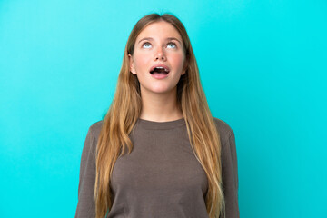Young blonde woman isolated on blue background looking up and with surprised expression